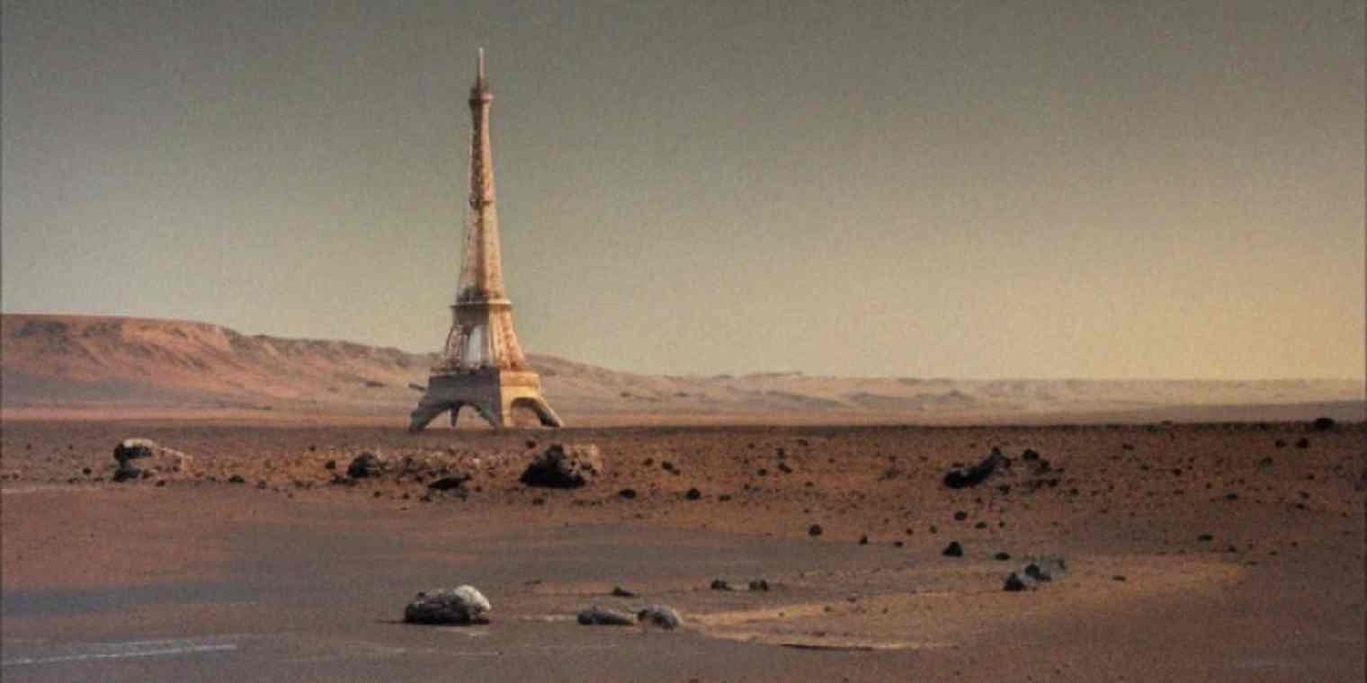 A surreal desert landscape resembling Mars with a solitary Eiffel Tower–like structure standing amid red sand, rocks, and distant barren hills under a muted sky.